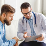 Medical practitioner in glasses showing test results to man and explaining diagnosis while sitting on couch in office of modern clinic