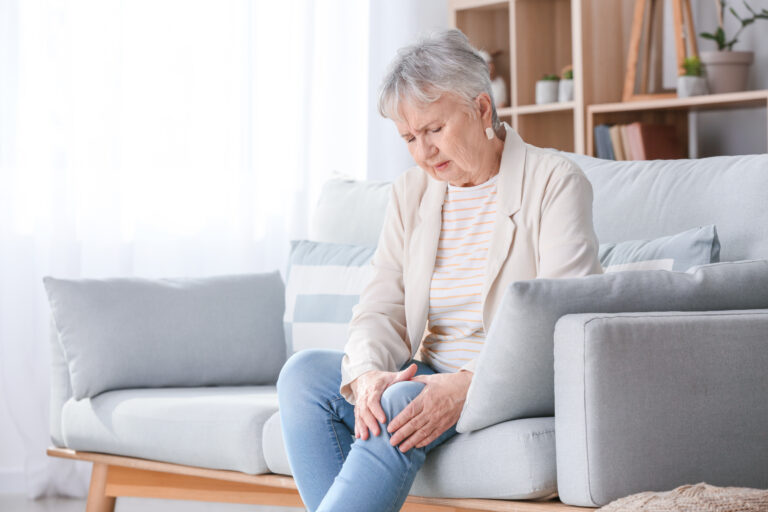 An elderly woman with short grey hair sits on a light grey sofa, looking down with a pained expression as she clutches her right knee with both hands. She is wearing a cream blazer over a striped shirt and blue jeans. The background is a bright, airy living room with a wooden bookshelf.