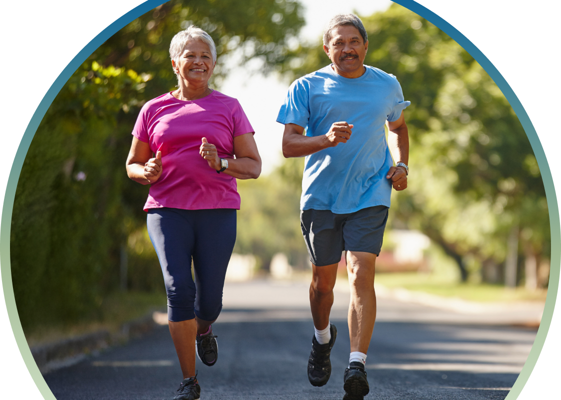 An active older couple jogs together down a sun-dappled, tree-lined street. The woman, on the left, wears a bright pink t-shirt and dark navy leggings, while the man, on the right, wears a light blue t-shirt and charcoal grey shorts. Both are smiling and mid-stride, framed within a large circular border with a soft green-to-blue gradient.