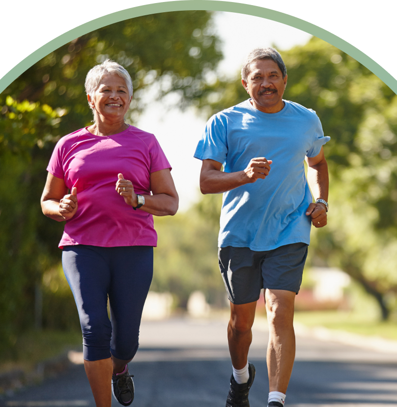 An active older couple jogs together down a sun-dappled, tree-lined street. The woman, on the left, wears a bright pink t-shirt and dark navy leggings, while the man, on the right, wears a light blue t-shirt and charcoal grey shorts. Both are smiling and mid-stride, framed within a large circular border with a soft green-to-blue gradient.