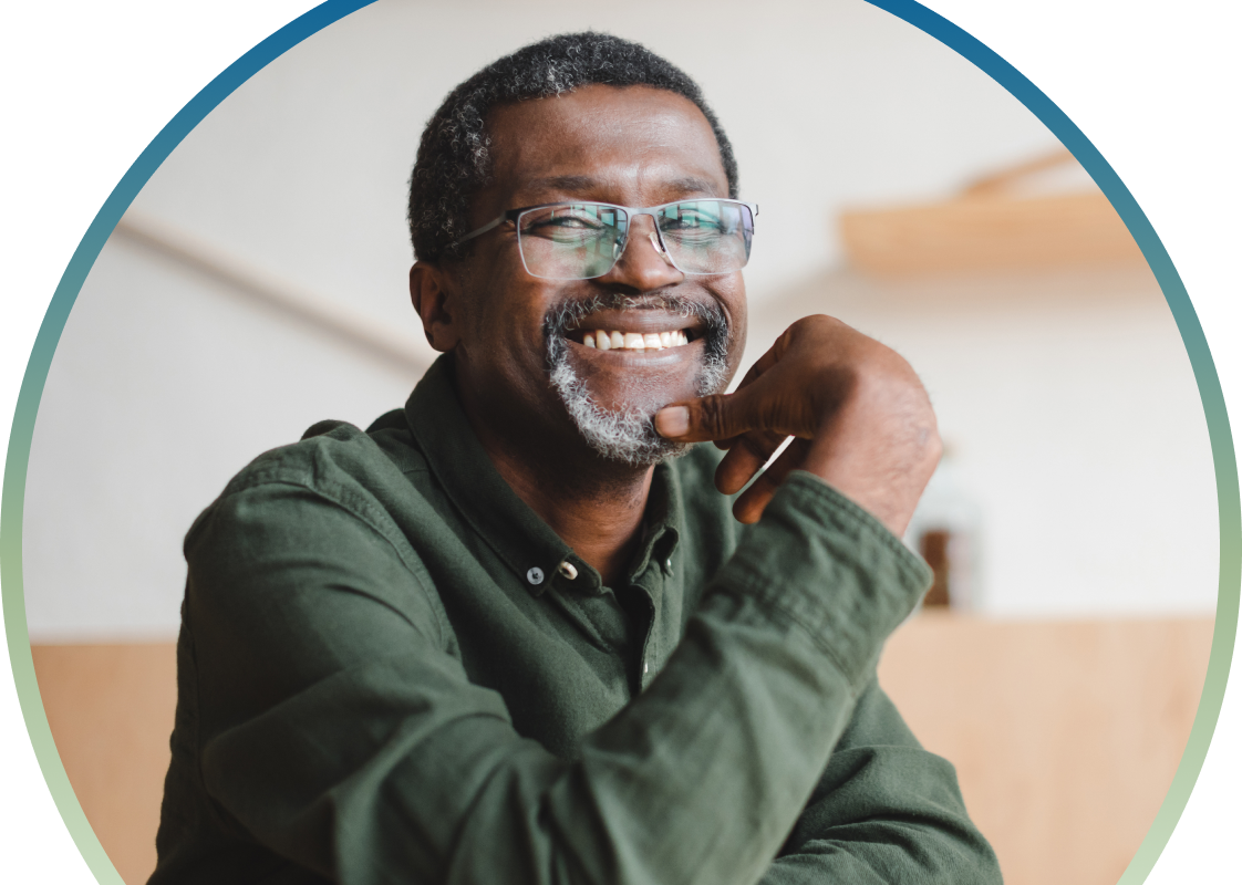 A smiling, middle-aged Black man with salt-and-pepper hair and a goatee wearing glasses and a dark green button-down shirt. He is resting his chin on his hand, looking off-camera with a thoughtful and happy expression. The image is framed within a circular teal border.