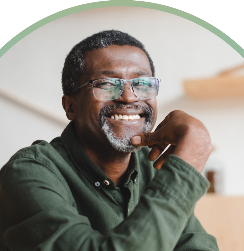 A smiling, middle-aged Black man with salt-and-pepper hair and a goatee wearing glasses and a dark green button-down shirt. He is resting his chin on his hand, looking off-camera with a thoughtful and happy expression. The image is framed within a circular teal border.