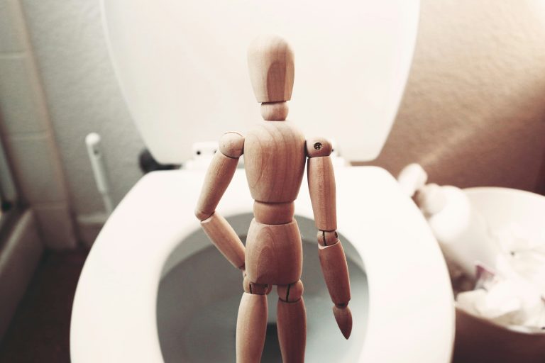 A wooden figure placed in front of an open toilet, suggesting the image of a man who may struggle to start peeing